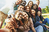 Elf junge Menschen unterschiedlicher Hautfarbe und Geschlechts, die auf einer Wiese sitzen und lachend ein Selfie machen