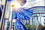Im Vordergrund vier Fahnenmasten mit im Wind wehenden EU-Flaggen, im Hintergrund das Gebäude des Europäischen Parlaments in Brüssel, darüber bewölkter Himmel (Quelle: Adobe Stock: Grecaud Paul)