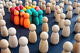 Mehrere bunte Holzspielfiguren stehen eng zusammen in einer Gruppe, rundherum holzfarbene, eher einzeln stehende Spielfiguren (Quelle AdobeStock: Vitalii Vodolazskyi)