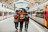 Drei Frauen stehen die Arme umeinander gelegt auf einem Bahnsteig zwischen zwei Zügen, dem Fotografierenden den Rücken zugewandt (Quelle: AdobeStock: lubero)